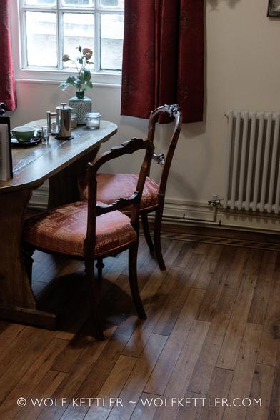 The photograph shows the inside of a cosy tea and coffee place. Main focus are two chairs and a table. In the background, top left, a window with red curtains.