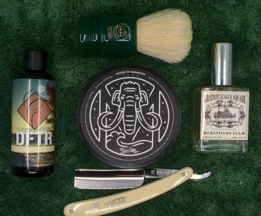 (ChatGPT description)

This image presents a premium wet shaving setup meticulously arranged on a dark green towel, featuring a selection of high-end grooming products and classic tools. At the center is a tub of shaving soap from House of Mammoth, labeled "Shire," showcasing the brand's signature mammoth logo in white on a black background. This artisan soap is known for its high-quality ingredients and unique scent profile.

To the left of the soap is a bottle of DFTR After Shave Lotion, which features a scenic label depicting a sunset over water, adding a rustic and natural aesthetic to the collection. On the right side, there's a bottle of Chatillon Lux Gratiot League Square Eau de Toilette, elegantly packaged in a clear glass bottle with vintage-style labeling, evoking a sense of old-world refinement and sophistication.

Above the soap is a Zenith shaving brush, notable for its dark green handle and natural boar bristles. The handle features the "2022 Brush of the Year" engraving, highlighting its special edition status and high-quality craftsmanship, making it a valued collector's item.

Below the soap is a Henckels Friodur 17 straight razor, featuring an ivory-colored handle and a stainless steel blade known for its durability and precision. The Henckels Friodur series is celebrated for its excellent edge retention and smooth shaving performance.

This thoughtfully curated setup embodies a perfect blend of tradition, luxury, and modern artisan products, ideal for wet s