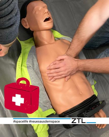 Eine Erste-Hilfe Übungspuppe, an der Herzdruckmassage geübt wird. Die Puppe ist hautfarben und liegt auf einem grauen Untergrund. Zwei Arme ragen rechts ins Bild, die Hände liegen auf dem Brustkorb der Puppe. Im Bild ist ein roter Erste-Hilfekoffer mit weißem Kreuz zu sehen.