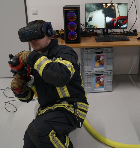 Feuerwehrmann in Hockhaltung mit VR Brille und Strahlrohr am Schlauch "im Anschlag".
Im Hintergrund steht ein illuminierter Gaming PC nebst Monitor, Tastatur und Maus auf einem Holztisch.
Auf dem Monitor ist eine geöffnete Tür zu einem verrauchtem Zimmer zu sehen, aus dem an der Decke Feuer schlägt.
Aus dem Stahlrohr im Bildschirm kommt ein Wasserstrahl der durch die Tür an die Zimmerdecke spritzt.