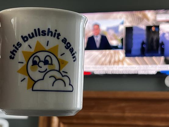 Coffee mug with a facetiously happy sun and the text “this bullshit again”. Out of focus news coverage of the inauguration proceedings in the background.