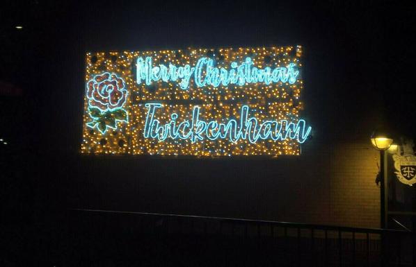 Lumières de Noël sur un mur de briques. À l'avant, il y a une rambarde métallique. À droite, il y a un lampadaire allumé et sa lumière est dans des teintes jaunes. Elle éclaire partiellement le mur ainsi qu'un panneau d'affichage sur lequel il y a des armoiries ou un blason. Ce blason est sur un fond blanc, La haut du blason est blanc et le bas et bleu. Dans le bleu,  il y a une croix blanche au milieu, et sur les quatre coin du blason, il y a, semble-t-il des oiseaus.
La lumière de Noël est un rectangle lumineux, dans des teintes orangées. Dans le haut il y a écrit : "Merry Christmas" et dans le bas "Twickenham".
À la droite du texte, il y a une fleur rouge avec des feuilles vertes. On dirait une rose, symbole de la ville ?