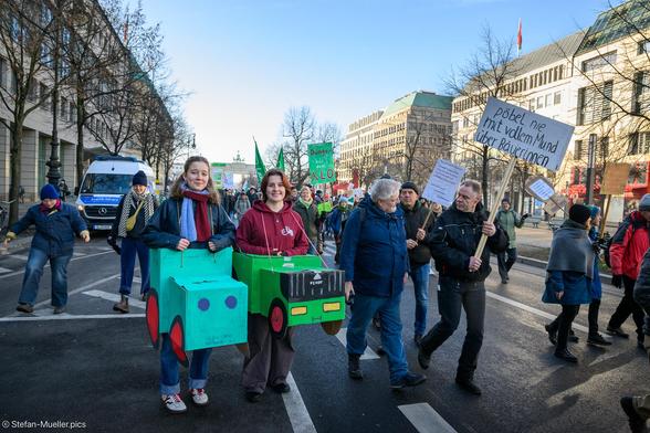 Demo-Teilnehmer*innen mit Papp-Treckern, da die richtigen Traktoren wegen der Maul-und-Klauen-Seuche nicht kommen konnten bei Wir haben es satt-Demo für eine andere Agrarpolitik. Bauer mit Schild: „Pöbel nie mit vollem Mund über Bäuerinnen“, Berlin, 18.01.2025