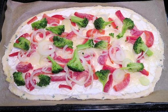 Das Bild zeigt einen Flammkuchen auf einem Backblech, belegt mit Creme, geriebenem Käse, roten Paprikastreifen, Zwiebelringen und kleinen Brokkoliröschen. Der Flammkuchen liegt auf Backpapier und ist noch ungebacken.