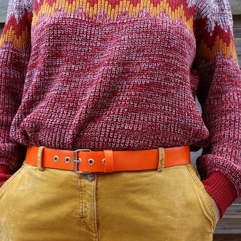 Ceinture orange en tuyau de lance à incendie upcyclée, portée par une modèle en pantalon de velours camel clair et gros pull en laine rouge, blanc et jaune