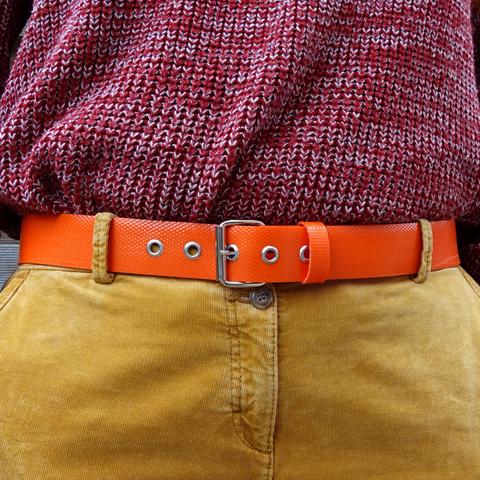 Ceinture orange en tuyau de lance à incendie upcyclée, portée par une modèle en pantalon de velours camel clair et gros pull en laine rouge et blanc