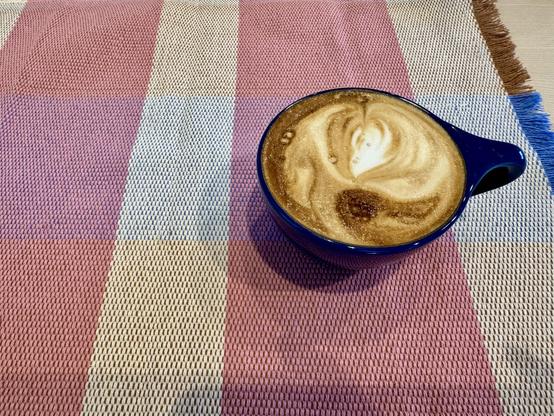 A small blue coffee cup filled with a frothy cappuccino sits on a blue, white, pink and brown placemat.