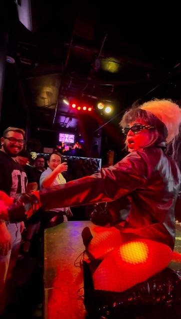 A drag queen in black thigh high boots, fishnet stocking, and leather jacket is performing on stage. She is on her knees arm stretched out to accept money being offered from the audience.  Different men, tall, short, Latino, Asian, can be seen near the edge of the stage watching and giving her money