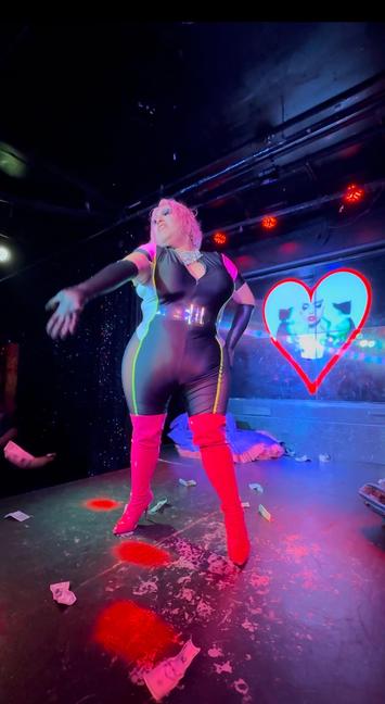 A drag queen in a black tight jumpsuit and red thigh high high heeled boots is onstage at a club arm out stretched with some dollar bills in the air.