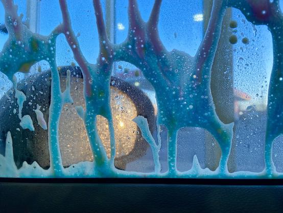 Looking out the passenger window in a car wash. The "tricolor" soap is dripping pink and blue down the windows, and the side mirror is reflecting the setting sun.