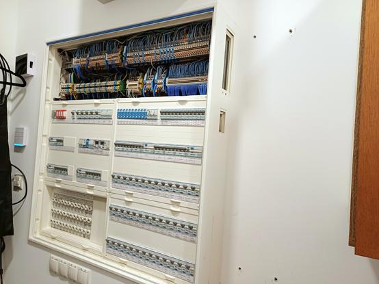 A single open electrical cabinet is shown on the left, containing circuit breakers and older remote dimmers. The side cover of the existing distribution board is removed, and drill holes in the wall are visible, marking the preparation for the installation of a new cabinet on the right. Highlighting the early stage of the installation process.