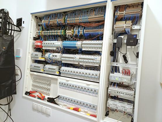 Two open electrical cabinets are shown. The left cabinet contains circuit breakers and remote dimmers (that are being replaced). The right cabinet is the new extension equipped with KNX components and the JUNG visualization server. Both cabinets have row terminals installed at the top for organizing wiring. Tools like screwdrivers and pliers are visible at the bottom.