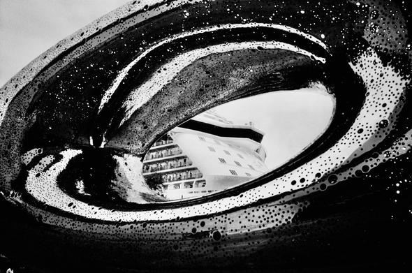 Black and white photography looking through a sculpture shaped like an oval donut.  The sculpture is polished metal and its surface is pierced with thousands of holes of different sizes.  Through the hole in the middle of the sculpture a cruise ship can be seen.
