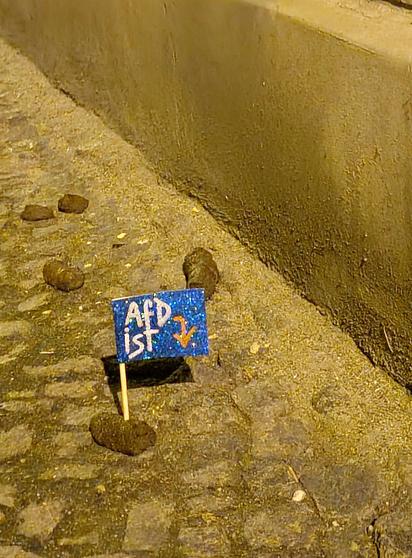 Photo eines Gehwegabschnitts an Hauswand, auf dem Boden verstreute Stücke Hundescheiße. Auf einem schönen dicken Stück ist eine kleine Flagge wie man sie sonst vielleicht auf Kuchen erwarten würde befestigt, mit der Aufschrift "AfD ist [und einem Pfeil nach unten in Richtung Kacke]"