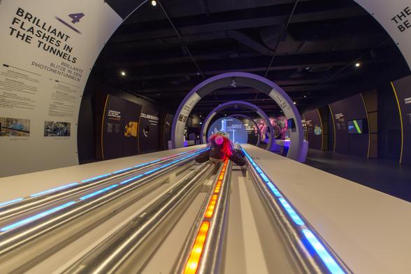 Max, das bunte Strickhorn, ist im Tunnelmodell des XFEL weitergekrabbelt, zu Position 4: Brillante Blitze in den Photonentunneln. Unter ihm weisen animierte Leucht-Pulse den Weg der Elektronen (rote, eine Strahlführung im Bild) und der Röntgenstrahlung (drei blaue Strahllinien). Eine Erklärtafel links im Bild gäbe ein paar weitere Informationen, ist aber tiefenunscharf abgebildet.