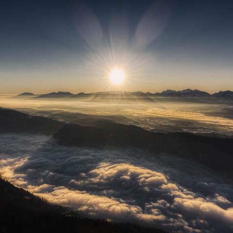 Sunrise above a mountain range, beneath them are forested hills, there is fog in the valleys