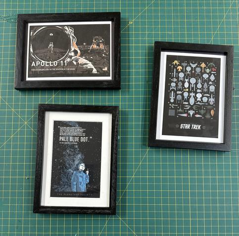 Three framed postcards on a green cutting mat. The top frame features an Apollo 11 theme, depicting an astronaut on the moon. The center frame displays the "Pale Blue Dot" concept with a person looking upward against a starry background. The third is a collection of Star Trek ships on a black background.  Cards from Chop Shop Studio.