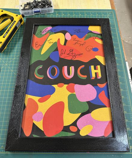 An ornate, colorful artwork framed in black, featuring an abstract design with various shapes in multiple colors. The center displays the word "COUCH" in bold, colorful letters, surrounded by signatures of different individuals. Tools and small hardware are visible in the background. Signed poster from Couch the band.