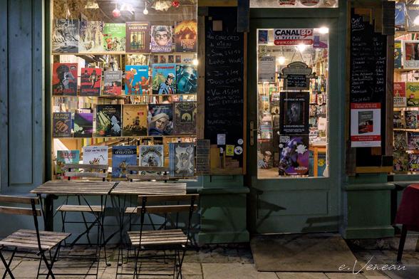Folding wooden tables and chairs in front of a bookstore window, lit up at night. Between the comic book covers, we can make out the bookseller’s face in profile.