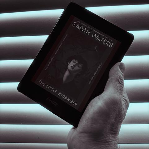 An e-reader displaying the cover of "The Little Stranger" by Sarah Waters, held in a hand against a backdrop of closed blinds with soft blue lighting.