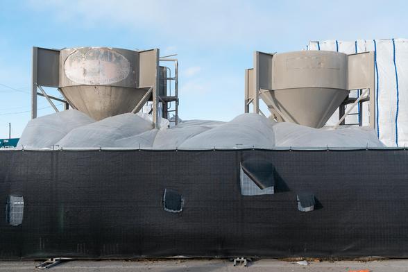 Two large metal objects stand behind a fence in a construction site under a creamy blue sky. The objects appear to be funnels supported by metal feet. The black burlap fencing contains numerous ripped square holes.