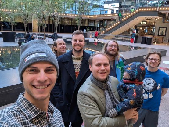 A group of people, including one holding a child, poses for a selfie in a modern indoor setting with trees and decorative lights. The atmosphere is casual and friendly, with others visible in the background.