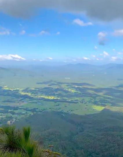 View from high above a valley, dotted with farms, trees and grassland. In the far distance, a rim of mountains in the afternoon haze melts into a blue blue sky dotted with tiny white clouds.