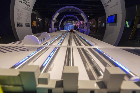 Max, das bunte Strickhorn, inmitten eines Modells des Tunnelsystems des Europäischen Röntgenlasers. Im Vordergrund sind die Gebäude des Forschungscampus Schenefeld zu sehen, dann die (unterirdischen) Tunnelröhren Richtung Hamburg. Diese sind teilweise erleuchtet, um die Pulse aus Röntgenstrahlen und Elektronen darzustellen. Ungefähr in der Mitte dieser Röhren, im gerade dunkeln Teil, hat sich unser Strickhorn "versteckt" und wartet darauf, gefunden zu werden.