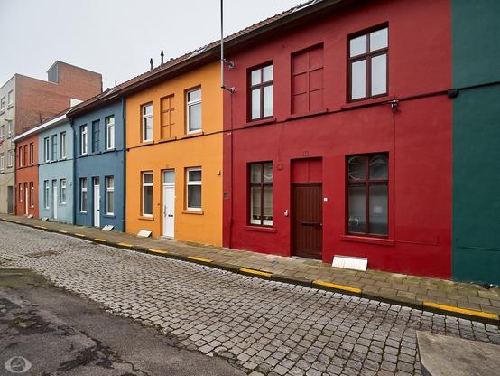 A line of coloured houses (from left to right: bright brown, cyan, navy blue, orange, red and dark green -the green house is barely visible) along an empty cobbled stone road.