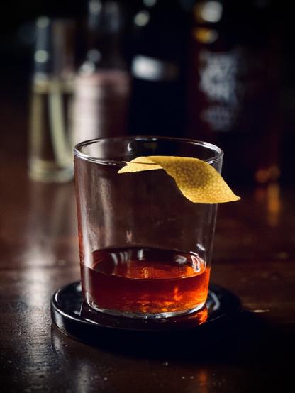 a dark red cocktail sitting neat in a glass with a lemon peel garnish with bottles of Sazerac rye whiskey, SC mai tai syrup, Peychauds bitters, and Pernod Absinthe in an atomizer bottle