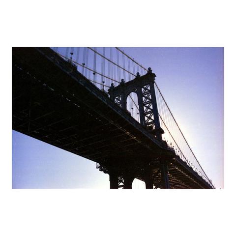 A photograph of the Manhattan Bridge in New York City. Shot from ground level, with the sun rising behind it. The sun reflection creates a warm glow under the bridge.