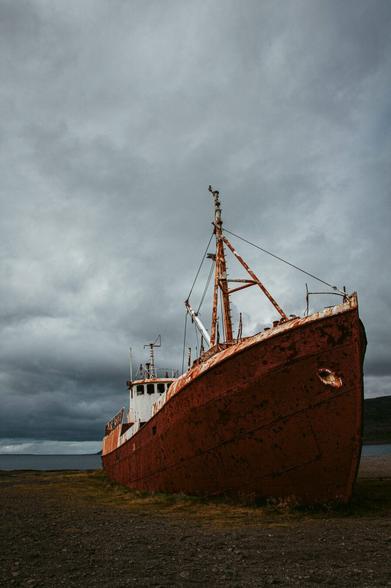 The image shows a rusted, decommissioned ship standing on solid ground, far from the water. The hull of the ship is a rusty red color, showing clear signs of weathering and decay. The sky is overcast and moody, creating a melancholic and atmospheric tone. In the background, a flat coastal landscape is visible, covered with grass and gravel. The scene conveys a sense of abandonment and tranquility.