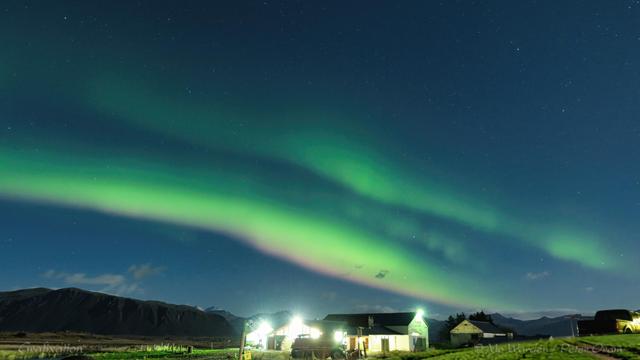 A colour photo of a night scene, with brightly lit farm buildings under a starry sky. Crossing the shot diagonally from lower right to centre left is a pair of auroral bands with some smaller ripples between them. The lower arc of the two has a distinct pinkish base, merging into the green above it.
