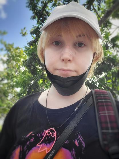 Selfie of Revy 
Wearing a white cap 
Her hair is short blonde
A black mask is under her chin 
She's wearing a i saw the tv glow necklace 
A plaid backpack arm is visible and slightly a purple and orange shirt of The Dude
