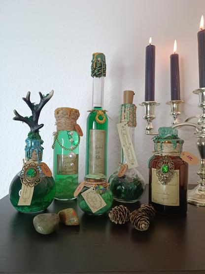 Sechs Flaschen stehen auf einer Holzoberfläche. Vier der Flaschen sind mit grünen Flüssigkeiten gefüllt und mit Perlen dekoriert. Eine Flasche ist braun und auf dem Deckel ist ein Edelstein angebracht. Eine Flasche ist mit Moos gefüllt und einer Lichterkette beleuchtet. Rechts im Hintergrund steht ein Entzündeter Kerzenleuchter. Im Vordergrund liegen zwei Kristalle und Tannenzapfen.