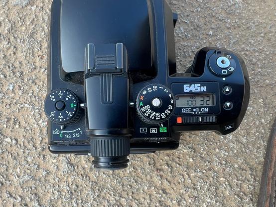 A top down view of a Pentax 645n, sitting on the ground. The lcd display shows that the camera is on shot 32, which is only possible because it is loaded with 220 film and is a 6x4.5 camera.