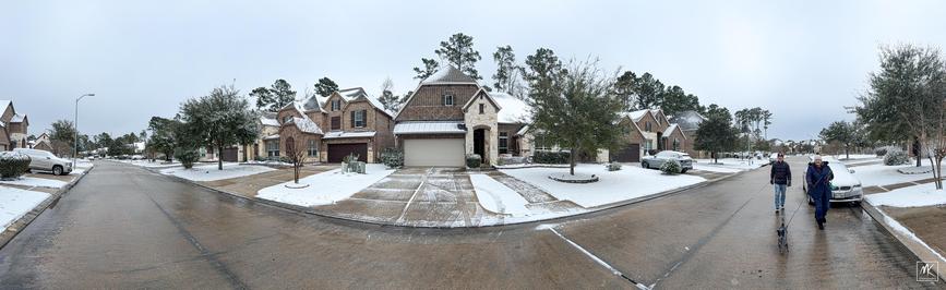 Color panoramic photo of a suburban street with snow on the houses and lawns.