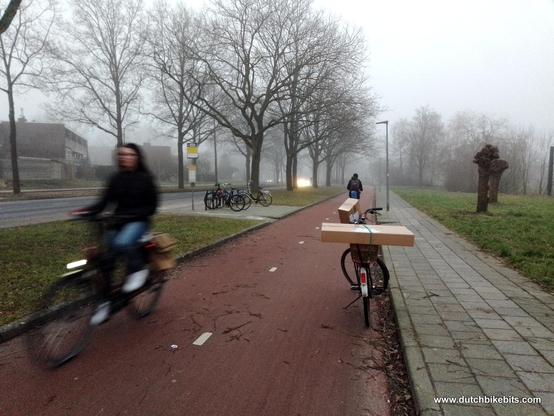 My bicycle parked on a Dutch cycle-path with parcels stacked on top. Cyclists heading in both directions past me. Cars on the other side of the wide verge, which also serves as cycle parking for the bus stop.