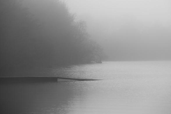 A black and white photograph of a muted and misty loch scene where a small jetty extends onto a clam water.