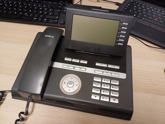 A Siemens Unify OpenStage 40 HFA VoIP phone is lying on a desk. It has a black casing with a handset on the left and a display on the top right. Below it are function keys as well as a keypad for numbers and navigation. In the background, a black keyboard and part of a laptop are visible.