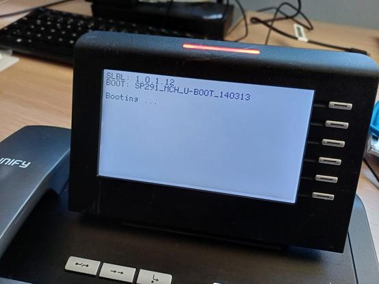 Close-up of the display of a Siemens Unify OpenStage 40 HFA VoIP phone during the startup process. The display shows:

"SLBL: 1.0.1.12
BOOT: SP291_MCH_U-Boot_140313
Booting..."

A red LED above the display is active. In the background, a keyboard can be seen out of focus.