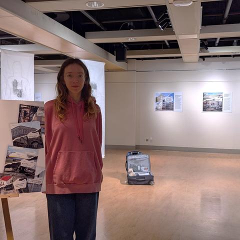 C'est une gallerie où se tient une exposition. Au plafond on voit des conduites d'aération et la structure, le sol est couleur bois, les murs sont blancs. Une jeune femme se tient debout, à côté d'un chevalet où figure une oeuvre de l'exposition. En arrière, au milieu de la pièce, on aperçoit une valise ouverte. Sur les murs, d'autres oeuvres sont accrochées,  de l'éclairage blanc est dirigé sur elles.