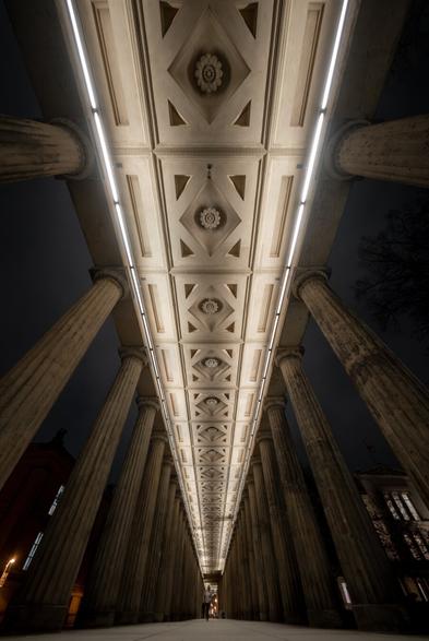 The image shows a symmetrical view of the colonnades on Museum Island in Berlin, captured at night. The perspective is directed upward, emphasizing the ornately designed ceiling of the colonnade with geometric patterns and floral rosettes. Long, rectangular light strips along the ceiling provide even illumination, highlighting the architectural details. Massive stone columns frame the scene on both sides. In the background, dimly lit facades of additional buildings are visible, while the silhouette of a tree is seen on the right. A single person stands in the distance under the colonnades, accentuating the depth of the architecture.
