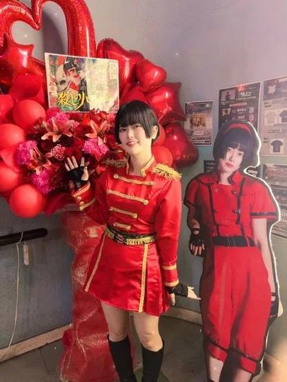 Chiyo in a plasic band uniform. She is infront of a large red flower stand and cardboard standee of herself in her Melon Betake outfig.
