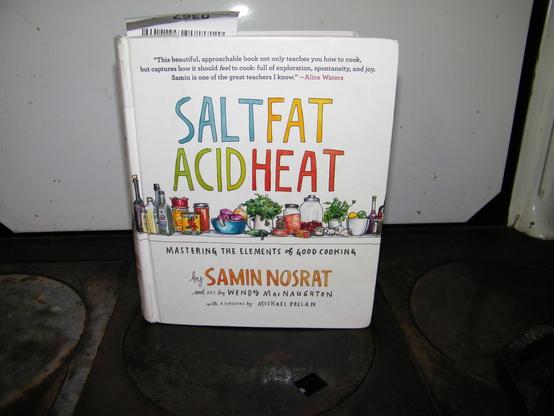 A cookbook on top of an old cookstove. The book's title is "Salt Fat Acid Heat: Mastering the Elements of Good Cooking" by Samin Nosrat and art by Wendy MacNaughton. Forward by Michael Pollan. The book cover is white, with colorful lettering and illustrations of food below the title.