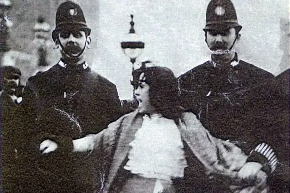 An old black and white photograph of Dora Thewlis, a young woman with dark hair, being held on either side by two policemen with identical bobby hats and moustaches. Dora has a fierce look on her face and is fighting the policemen. She was being arrested at a march on Parliament for women's suffrage in 1907.
