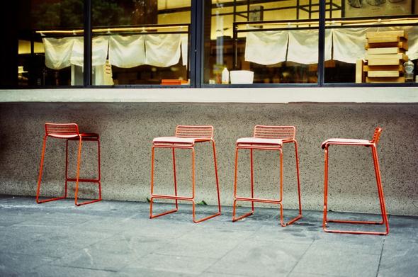 Photograph of a cafe window from the street outside.  The cafe is closed.  Outside are four orange steel stools on the footpath. Through the cafe window a stack of wooden food trays are visible, as well as small fabric curtains hanging from bamboo rods.
