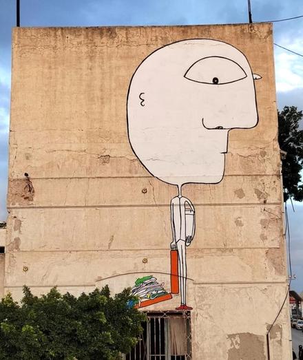 Streetartwall. A large mural of a traveler has been painted on the beige exterior wall of an old multi-storey building. The mural is designed like a simple drawing and shows a bald man with a huge head. He has one large eye, a pronounced nose and a small line for a mouth. His body is very narrow and small, he wears a white suit and tiny red shoes. He has a kind of hatbox tucked under one arm and a suitcase in the other hand. The red suitcase with all his clothes has opened and his clothes are spread out on the floor behind him. An unusual and magnificent mural by a Tunisian artist and illustrator living in Paris.