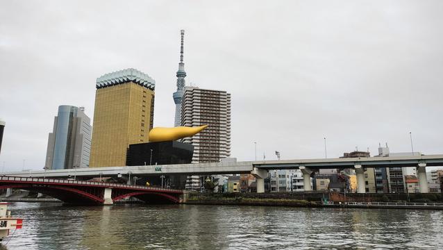 A river in Tokyo. On the far side are several buildings, both modern high rise buildings and older ones about four stories tall. One building resembles a black box with a golden elongated shape, pinched at one end, on top. A tall metal tower is visible behind the buildings.