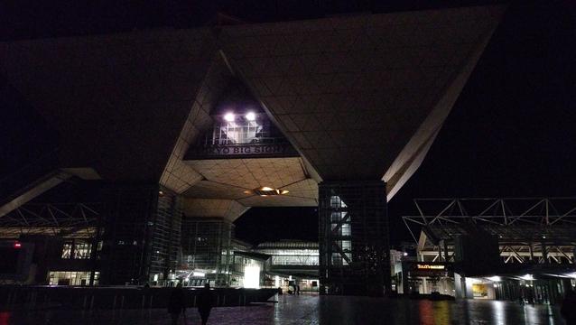 A night scene of a large plaza in Tokyo. The scene is dominated by an unusual modern building that resembles four upside down pyramids resting on glass and metal structures with staircases inside. There is a sign at the front of where the two front pyramids converge that reads TOKYO BIG SIGHT. It is 190 feet or 58 meters tall. Smaller structures and a few pedestrians are visible in the background.